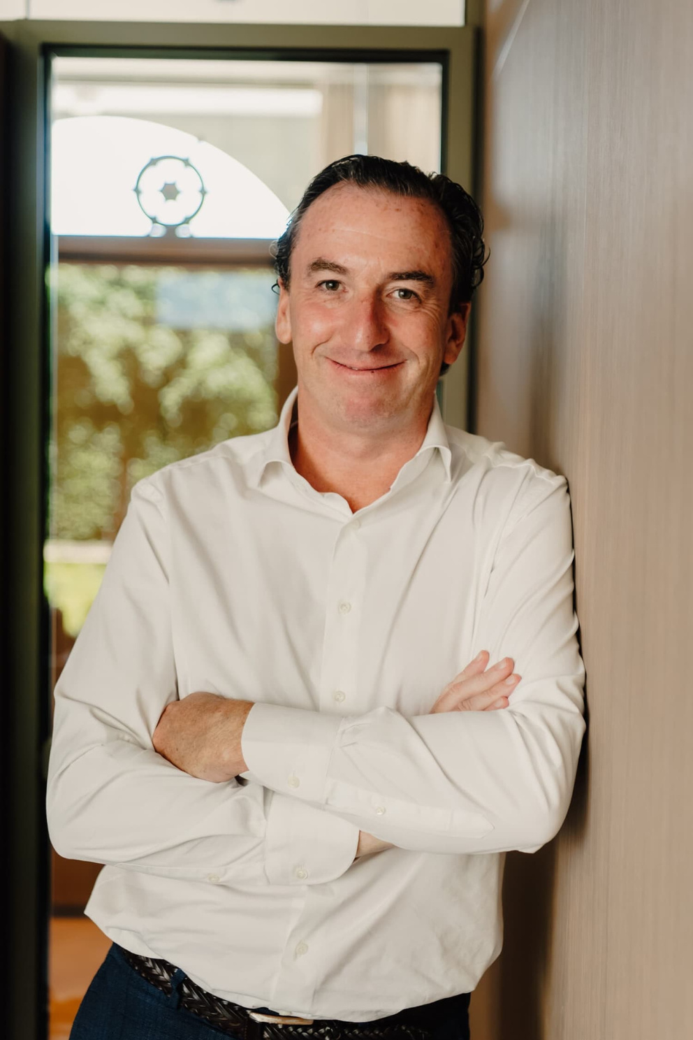 Man in witte blouse met opgestoken armen leunt tegen een muur, glimlachend, in een lichte, moderne omgeving.
