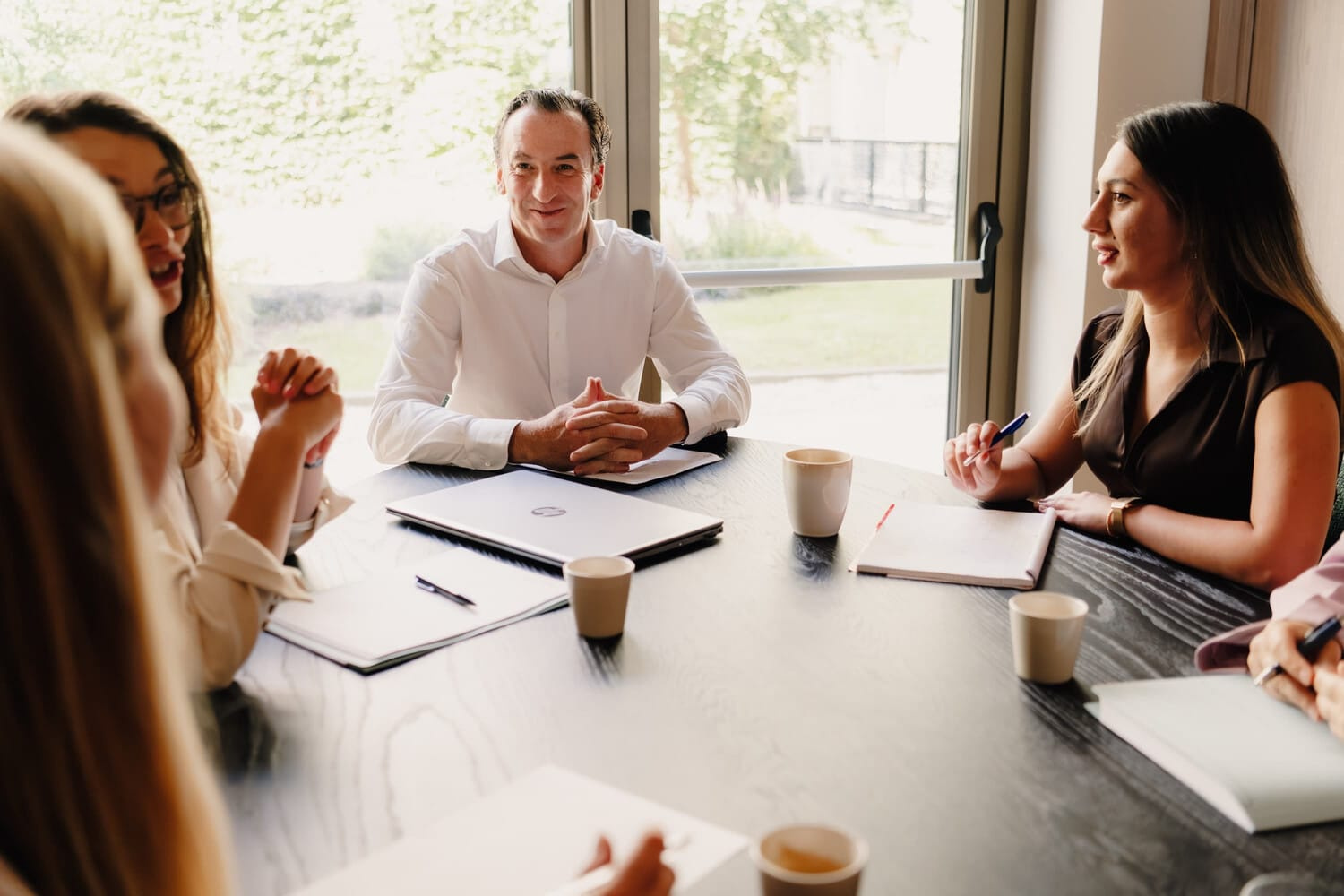 Een groep mensen in zakelijke kleding vergadert rond een tafel, met notitieboekjes en koffiekopjes in zicht.