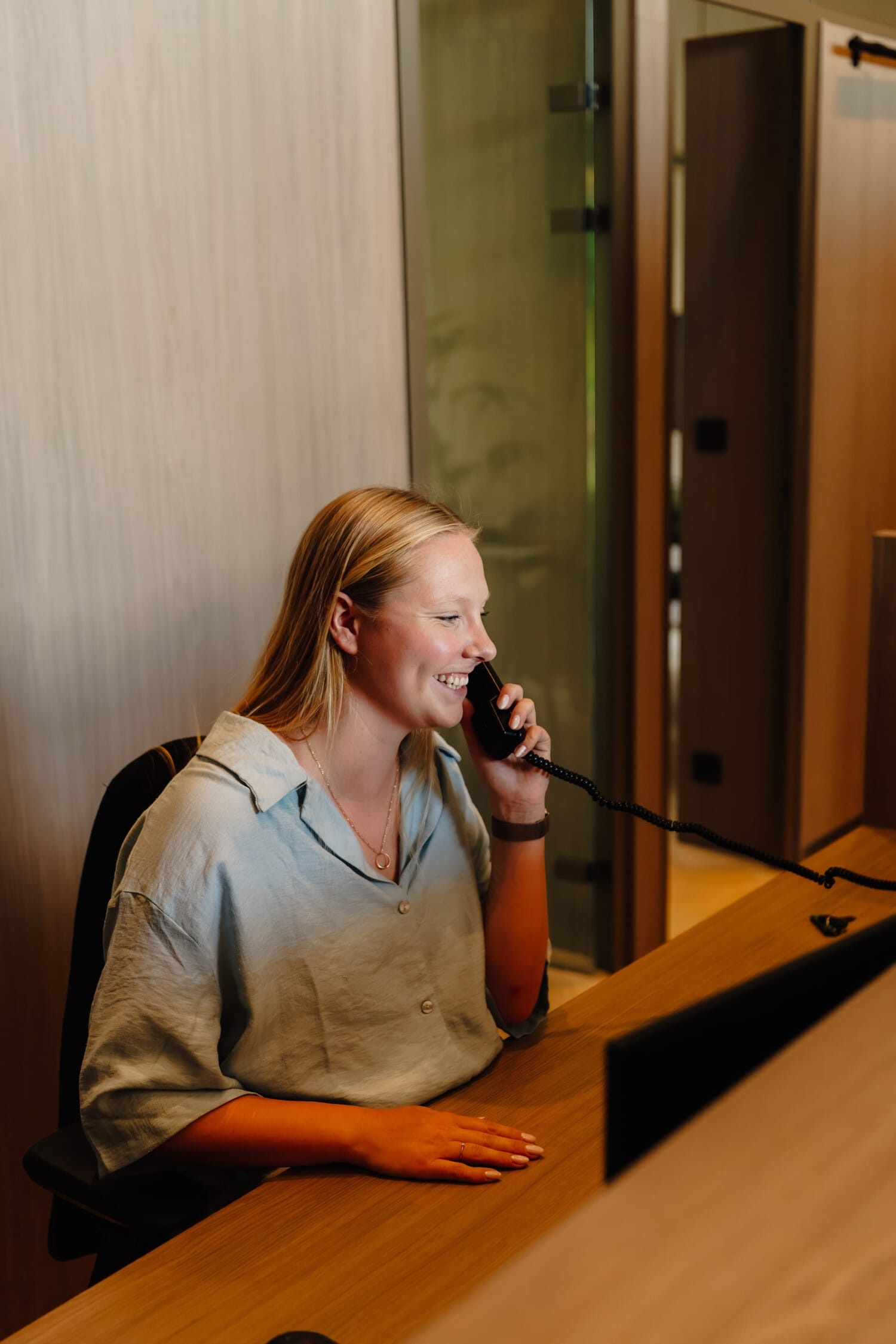 Blonde vrouw in een kantooromgeving, lacht terwijl ze een telefoongesprek voert en aan een bureau zit.
