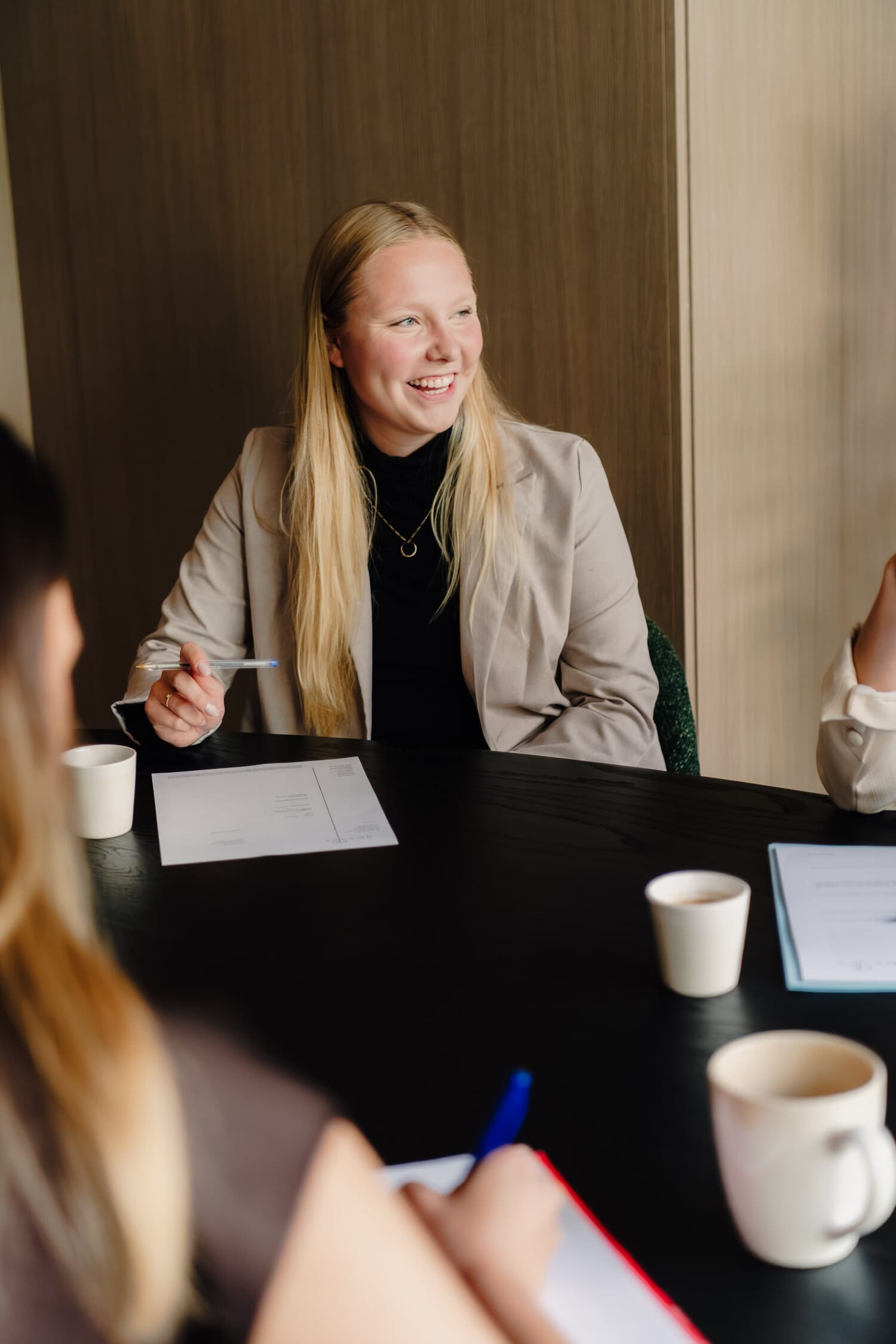 Een jonge vrouw met lang blond haar, glimlachend aan een vergadertafel, omringd door anderen.