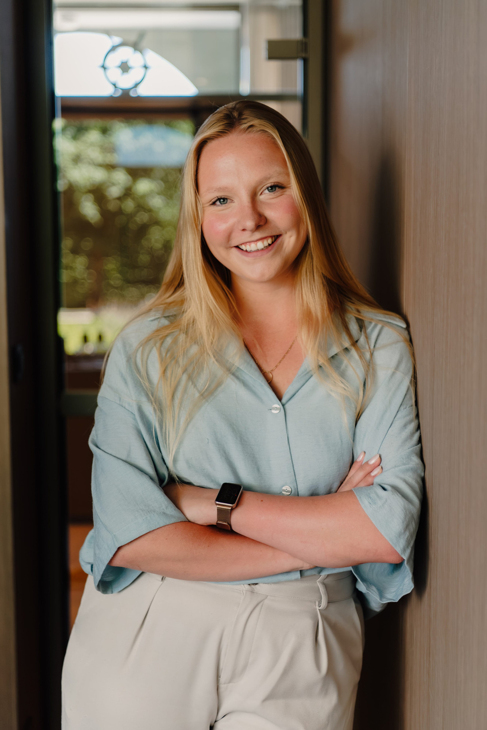Een lachende vrouw met lang, blond haar, gekleed in een lichtblauw shirt, leunt tegen een muur in een moderne binnenruimte.