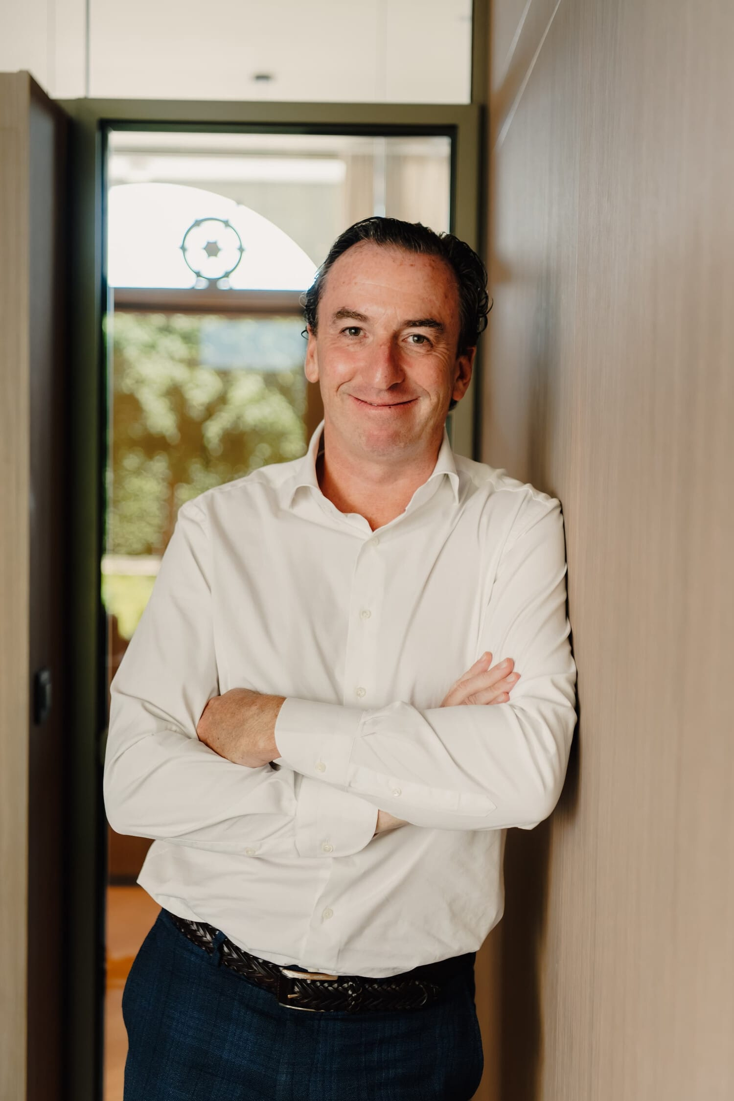 Man in een witte blouse leunt ontspannen tegen een muur met zijn armen over elkaar, in een moderne omgeving.