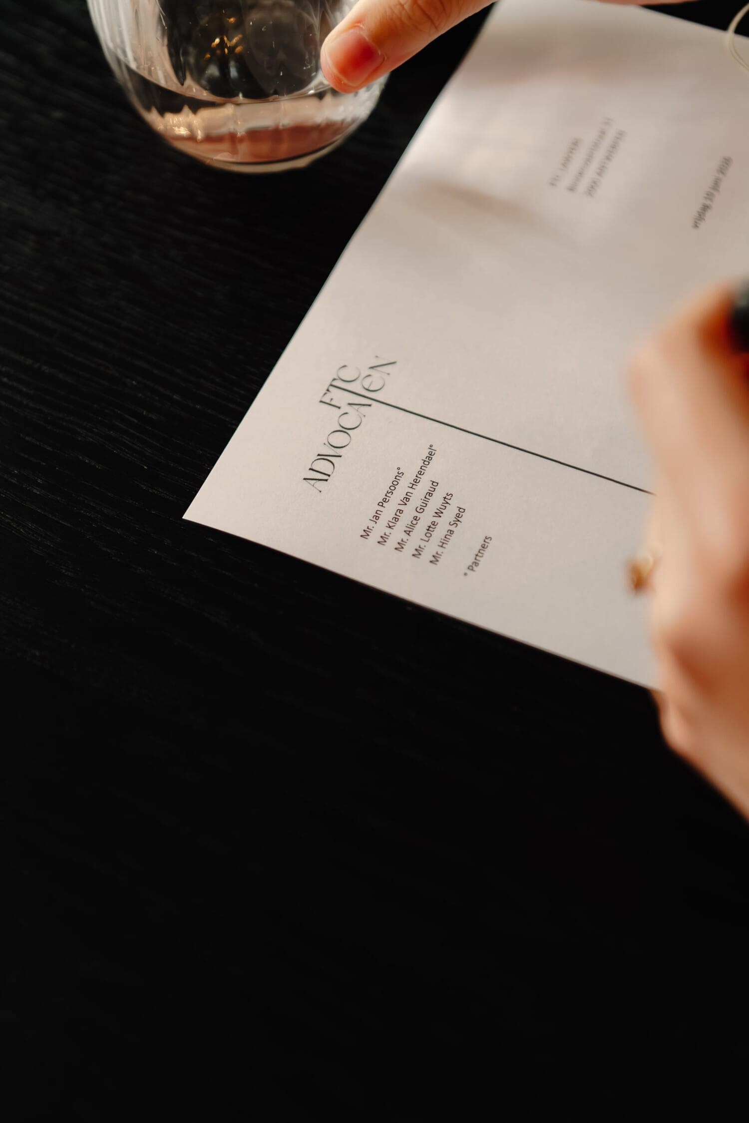 Een hand schrijft met een pen op een blanco brief met een elegante bovenkant en een glas water op de achtergrond.