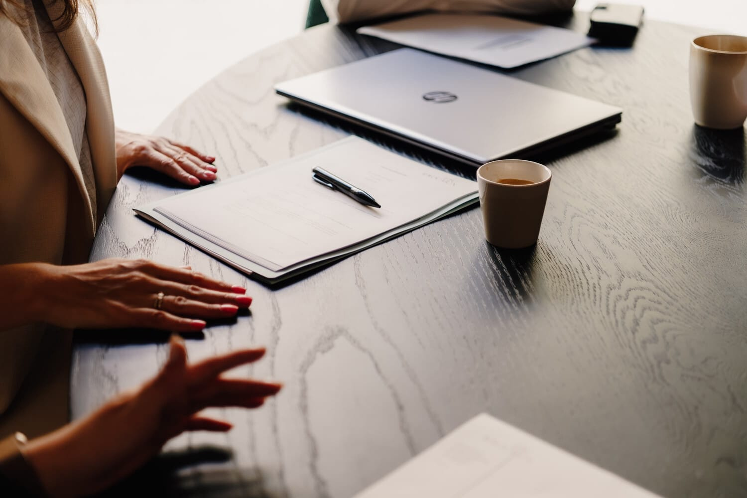 Een ronde tafel met notitieblokken, een laptop en koffiekopjes. Twee handen zijn zichtbaar met gekleurde nagels.