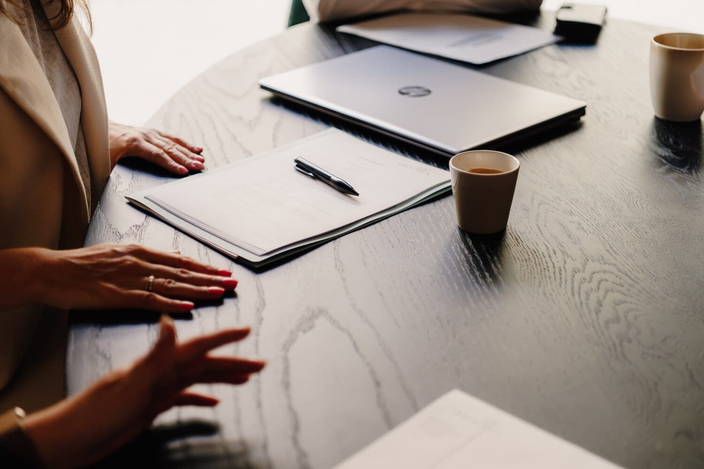 Een ronde tafel met notitieblokken, een laptop en koffiekopjes. Twee handen zijn zichtbaar met gekleurde nagels.