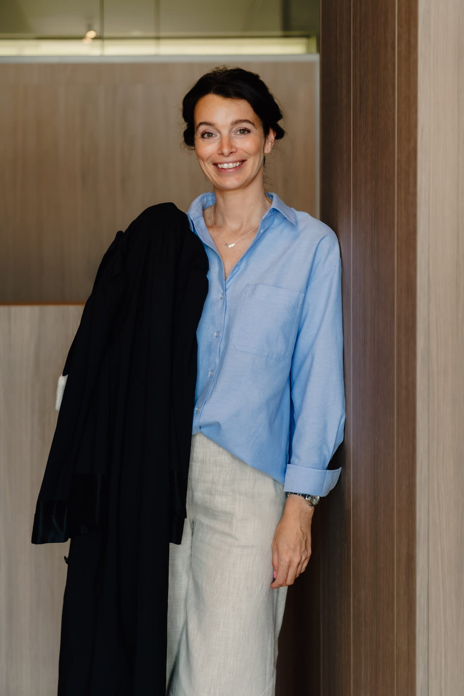 Een vrouw met een glimlach draagt een lichtblauw shirt en beige broek, leunend tegen een wand van hout.