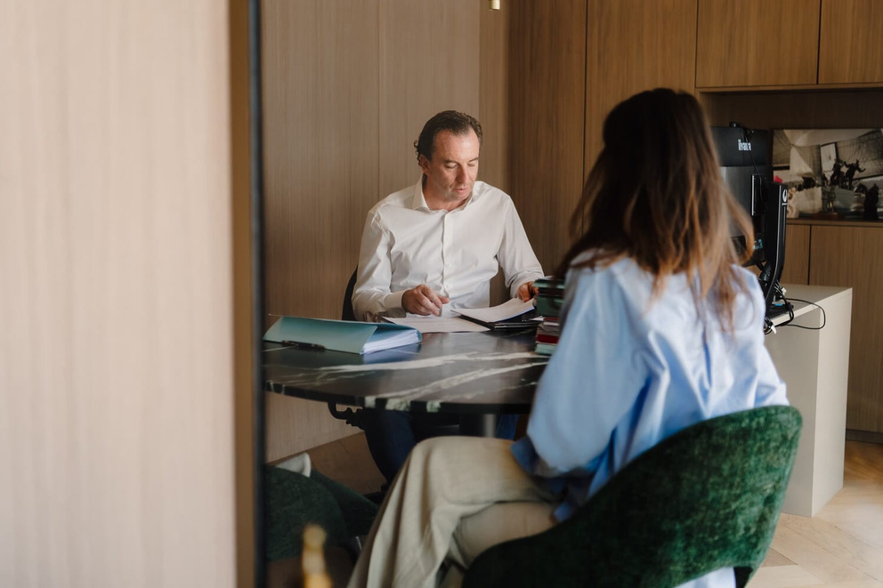 Man met papieren zit aan een tafel, terwijl een vrouw in een blauwe blouse naar hem kijkt. moderne kantooromgeving.