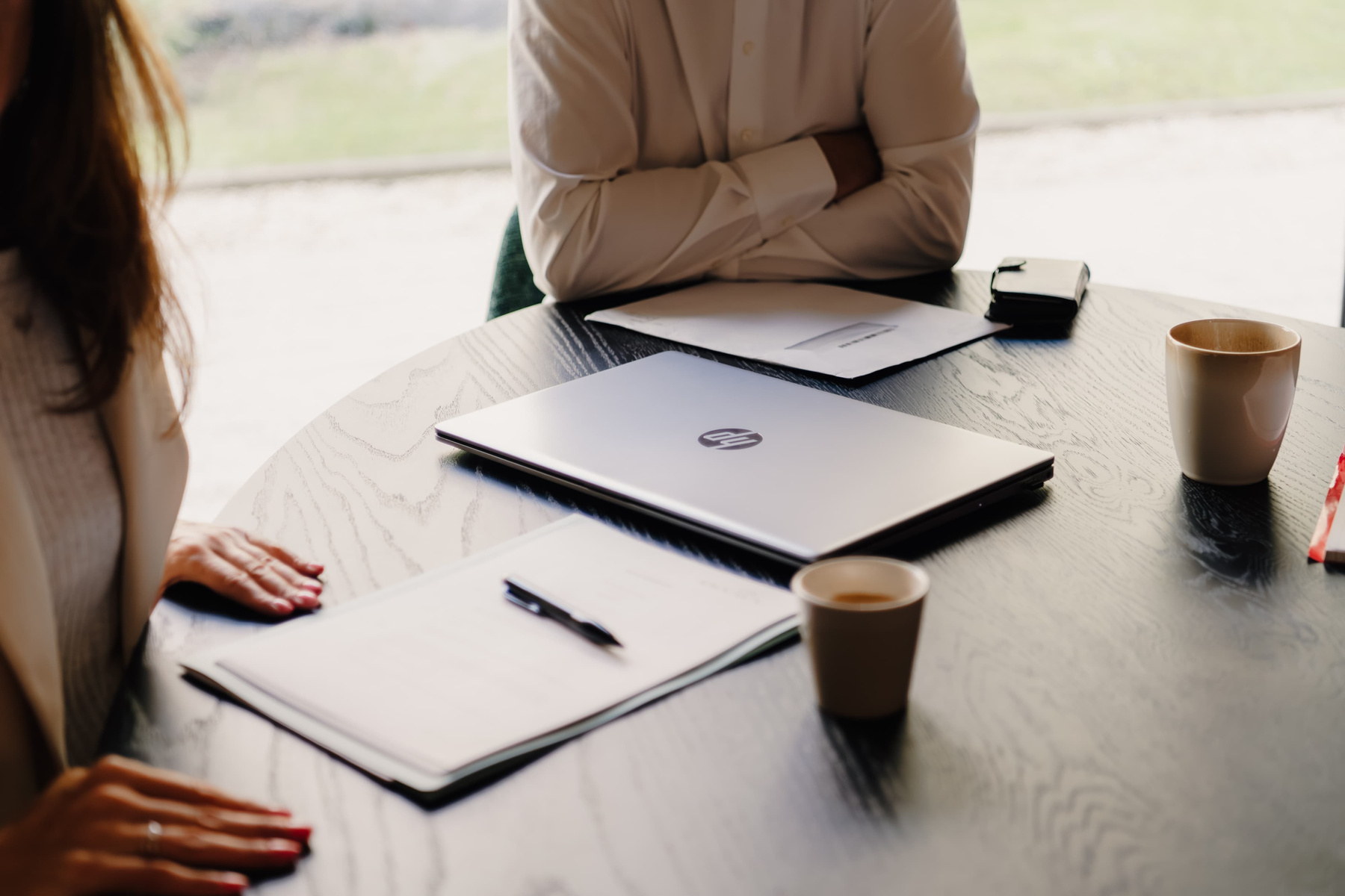 Een ronde tafel met een laptop, papieren, een pen en twee koffiekopjes, met handen van personen zichtbaar.