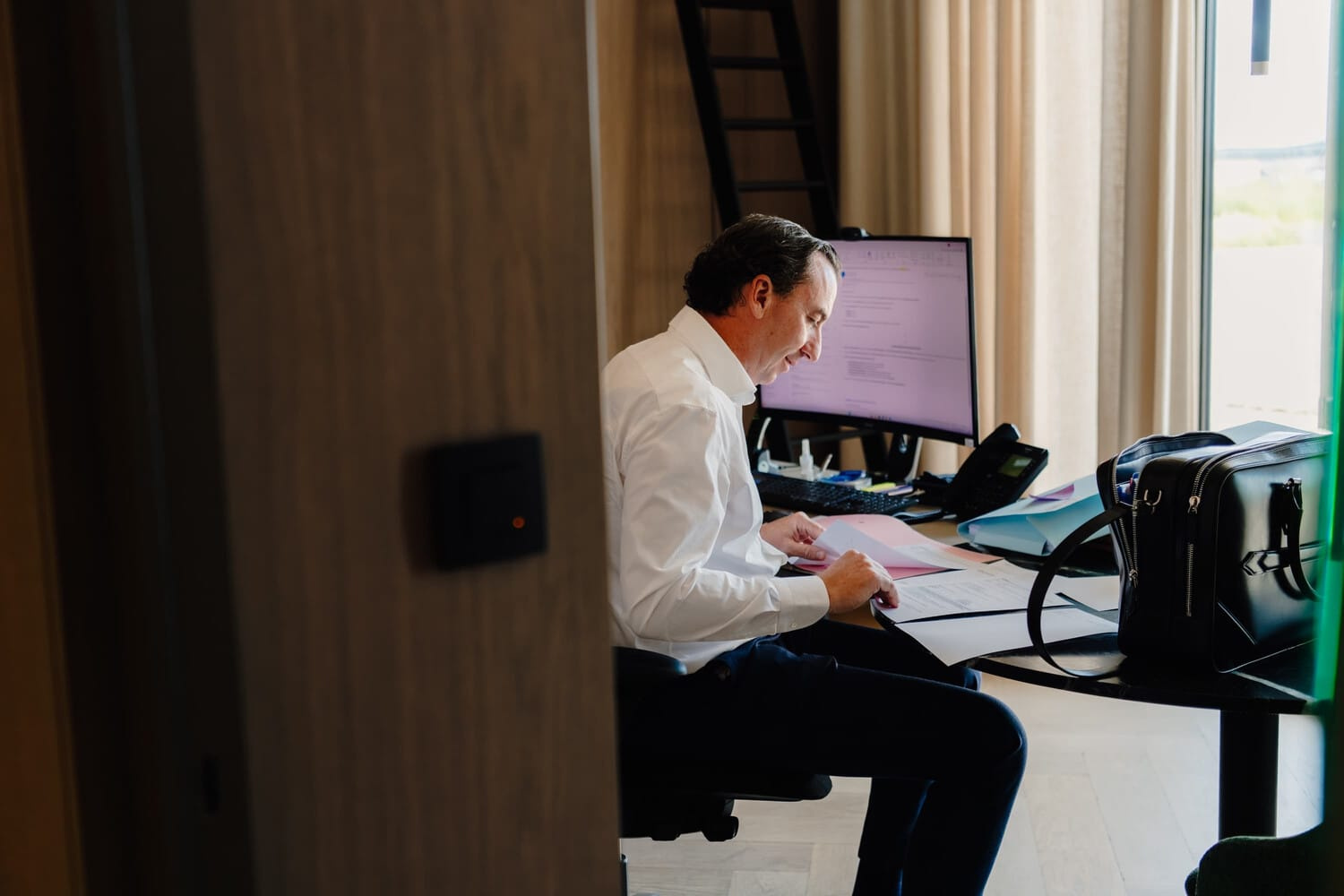 Een man in een wit shirt zit achter een bureau en werkt geconcentreerd op documenten naast een computer.