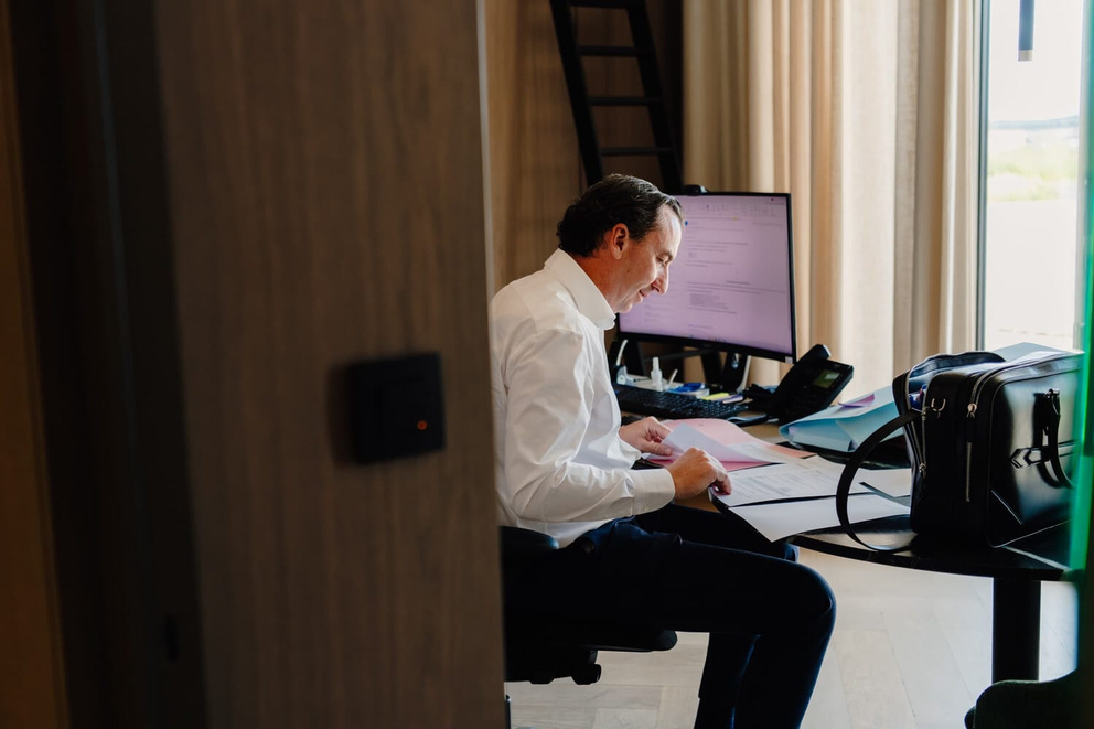 Een man in een wit shirt zit achter een bureau en werkt geconcentreerd op documenten naast een computer.