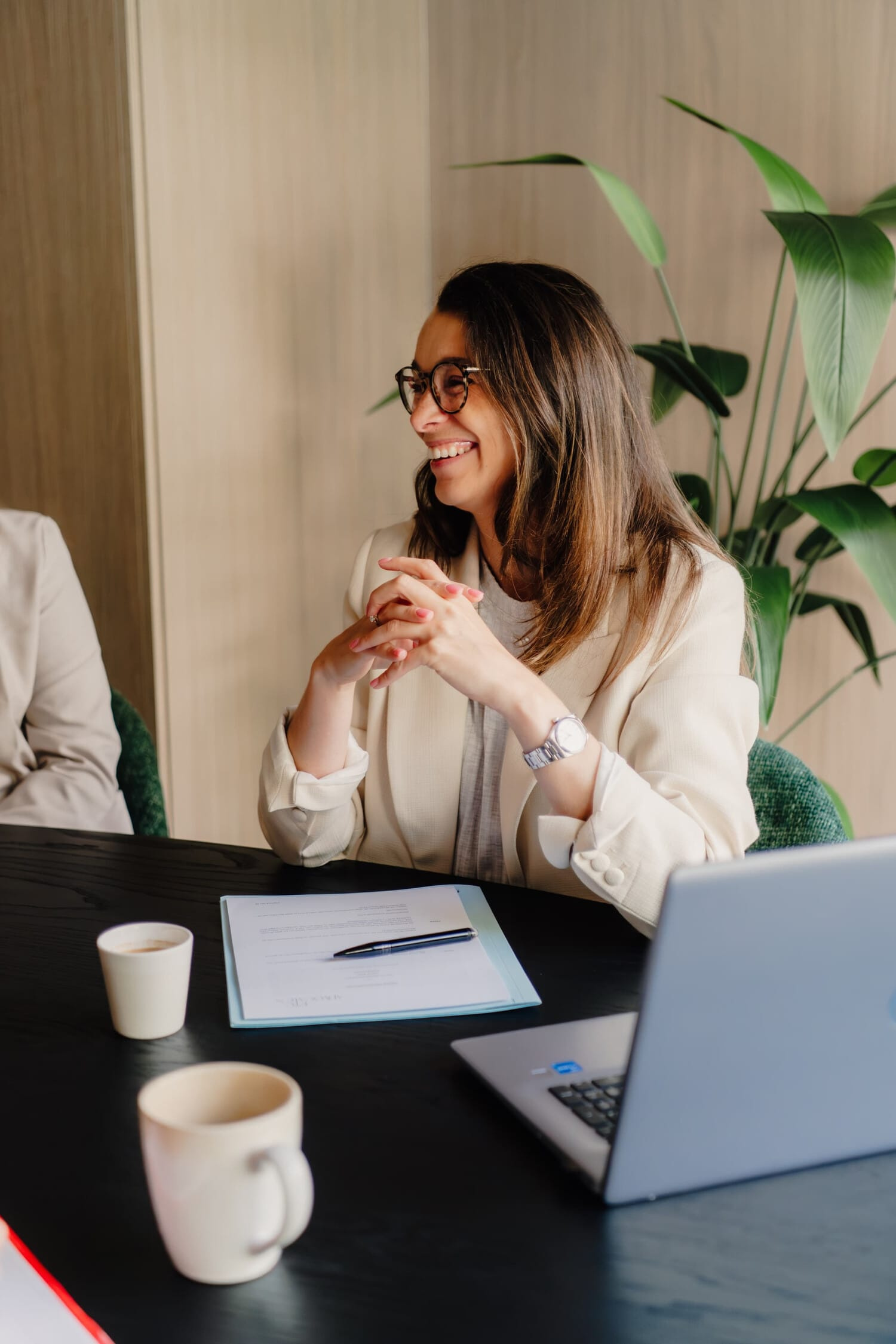 Vrouw in zakelijke kleding lacht tijdens een gesprek aan een tafel, met planten en een laptop op de achtergrond.