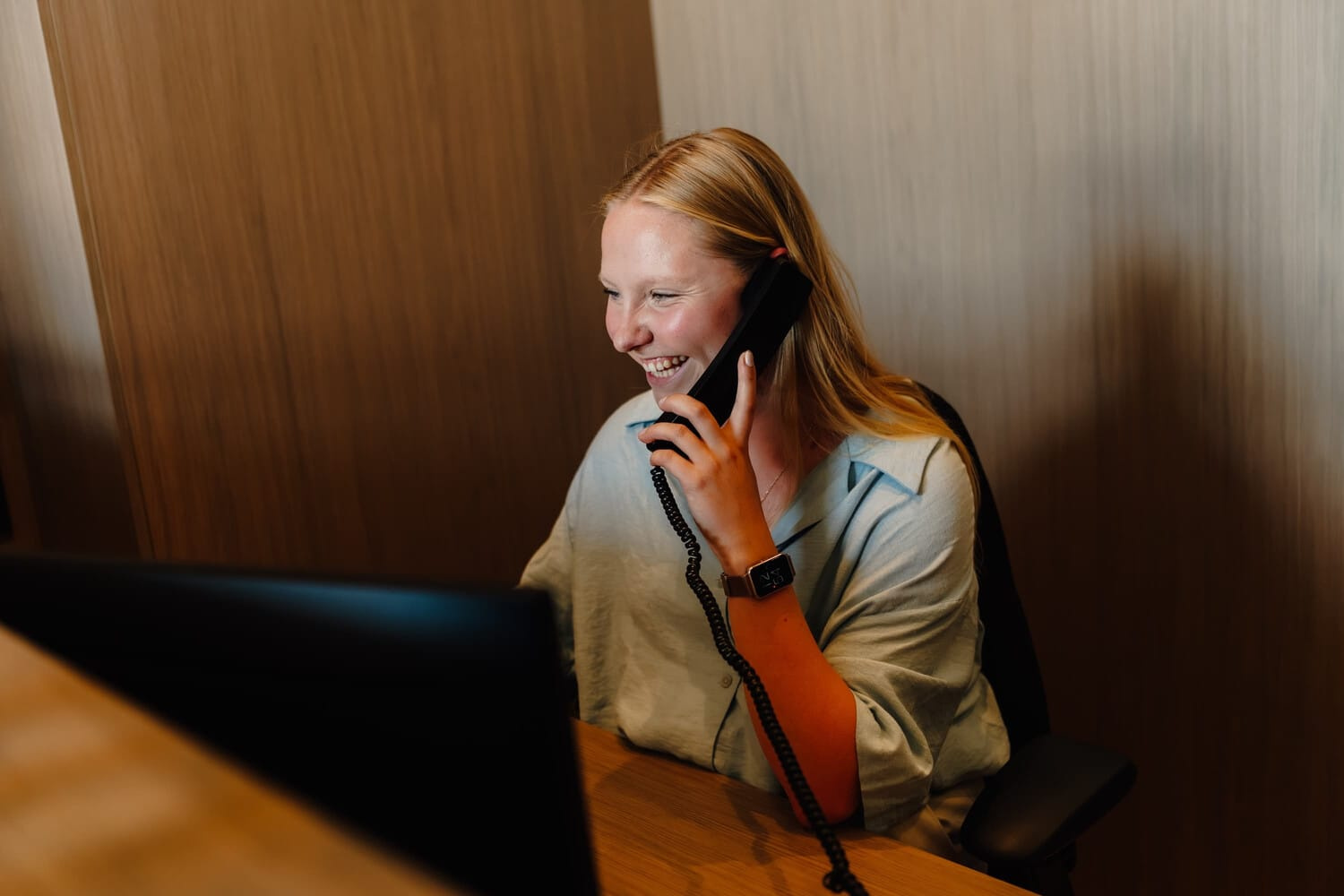 Een lachende vrouw met lang blond haar spreekt telefonisch terwijl ze aan een bureau zit.